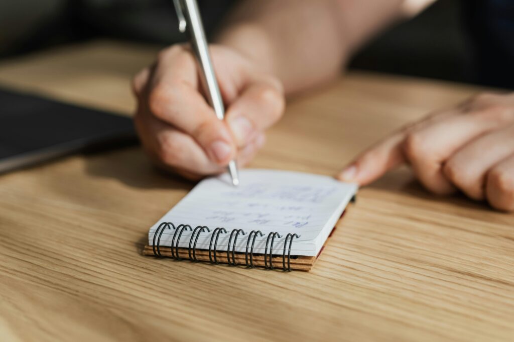 From above of crop faceless male student writing with pen in notebook at wooden desk during lesson